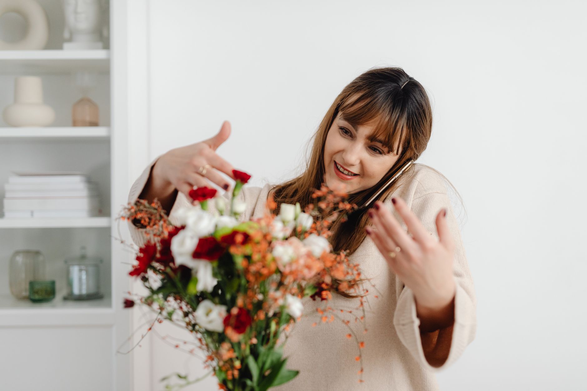 Smiling woman multitasking, arranging bouquet while talking on smartphone in cozy room.