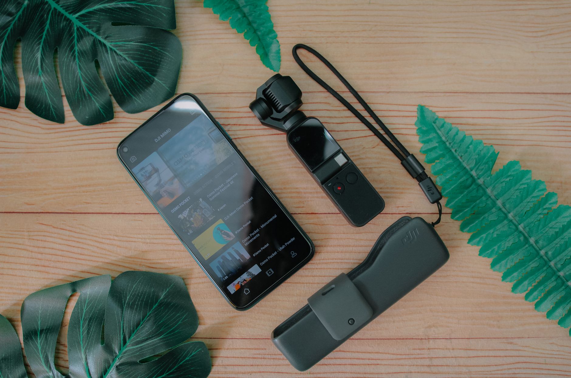 A flat lay of a smartphone and camera with green leaves on a wooden table.