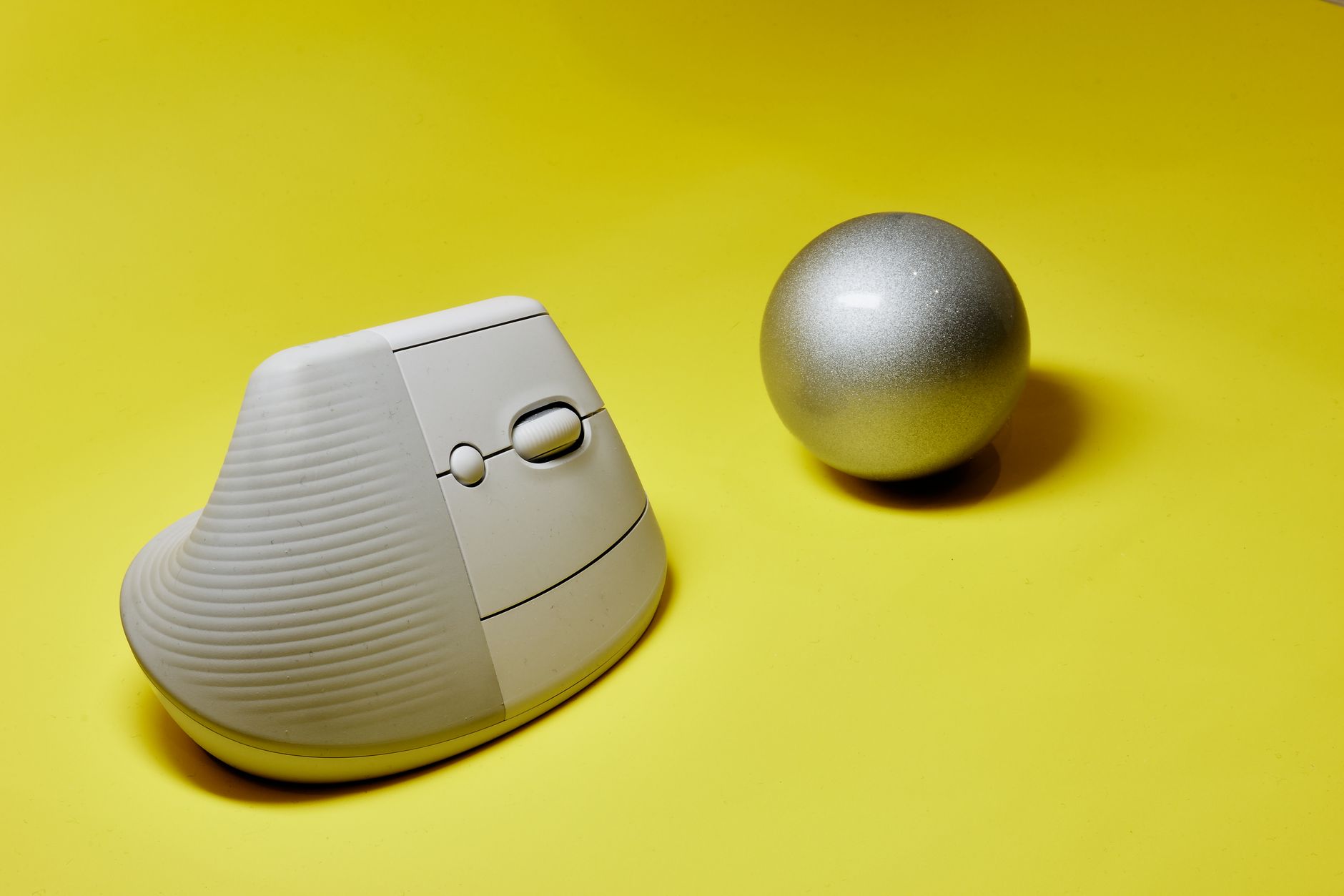 A sleek ergonomic vertical mouse next to a metallic sphere on a vibrant yellow background.