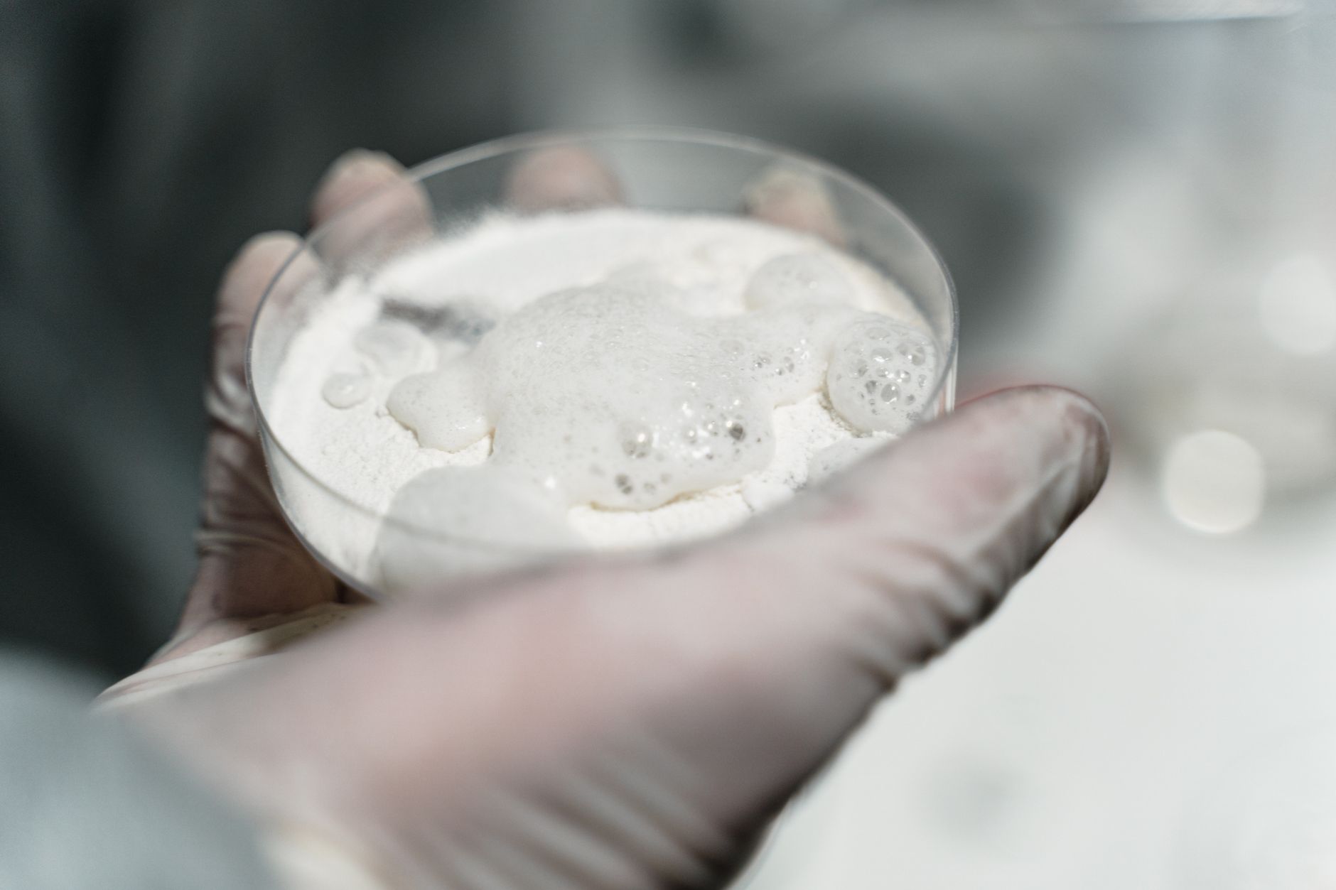 Close-up of a gloved hand in a lab holding a petri dish with a foamy substance, possibly for scientific analysis.