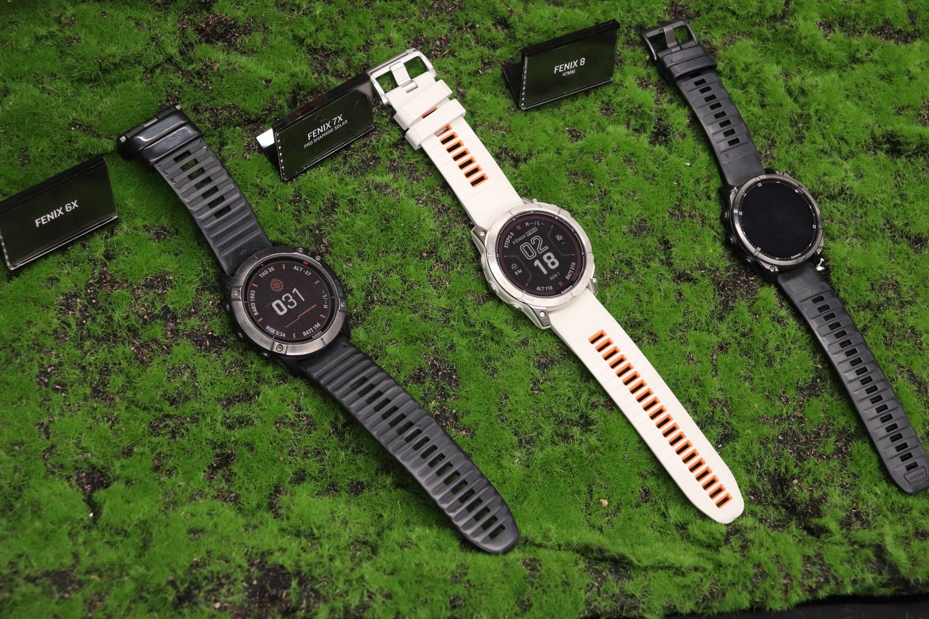Three outdoor watches displayed on a textured green surface, featuring Fenix model options.
