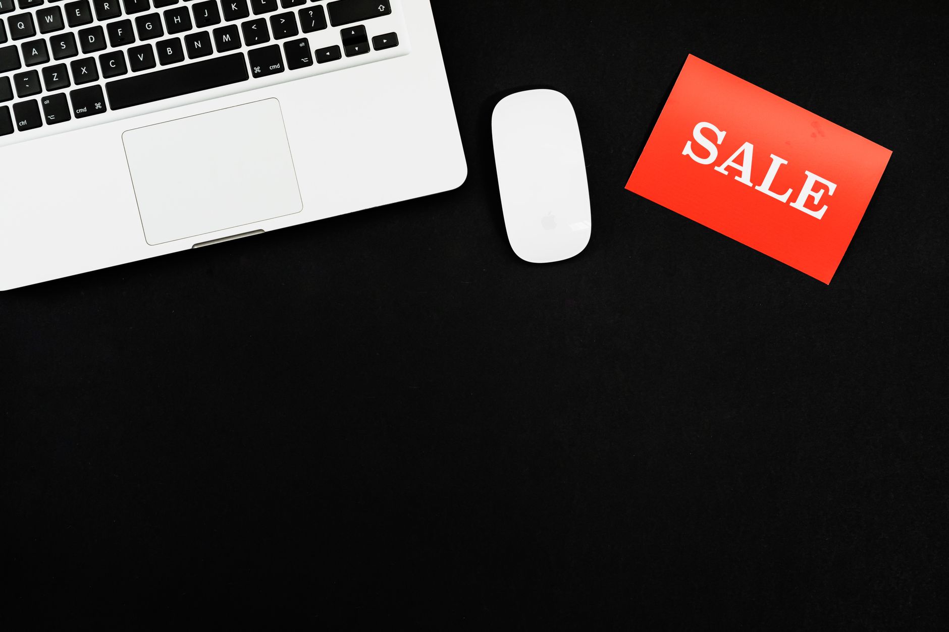 Minimalist flat lay of a laptop, mouse, and a red sale sign on black background.