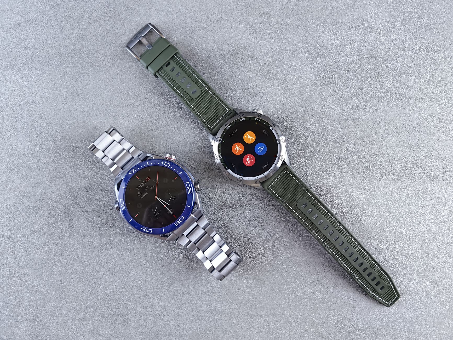 Close-up of two smartwatches with metallic and green straps on a textured concrete background.