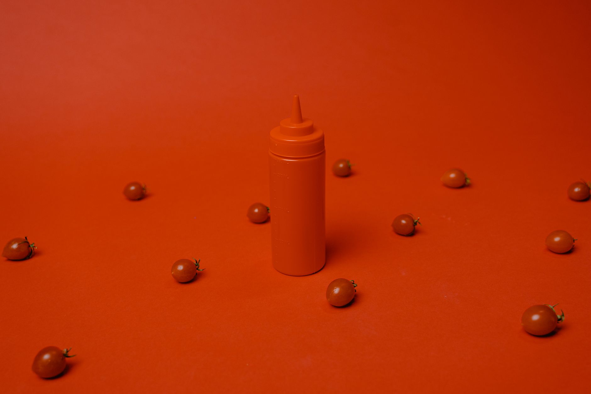 Red bottle of ketchup surrounded by cherry tomatoes on red background. Simple and bold.